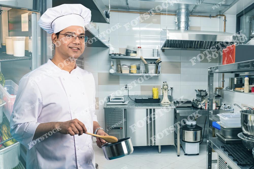 Повар в белой униформе и кухонной шапке стоит на профессиональной кухне. Он держит в руках кастрюлю и кухонную лопатку, улыбается, смотрит прямо на камеру. На заднем плане видны кухонное оборудование, столешницы и различные кухонные принадлежности.